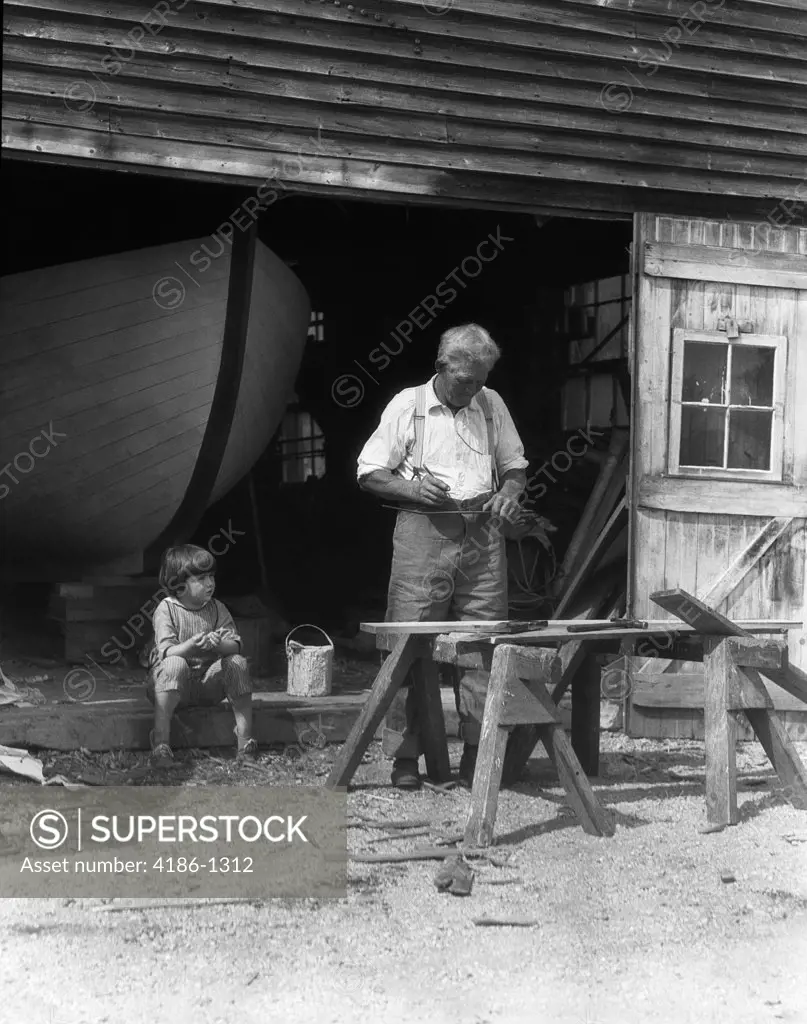1920S 1930S Elderly Man Grandfather Working At Saw Horses As Little Boy Watches Hull Of Boat Visible In Background