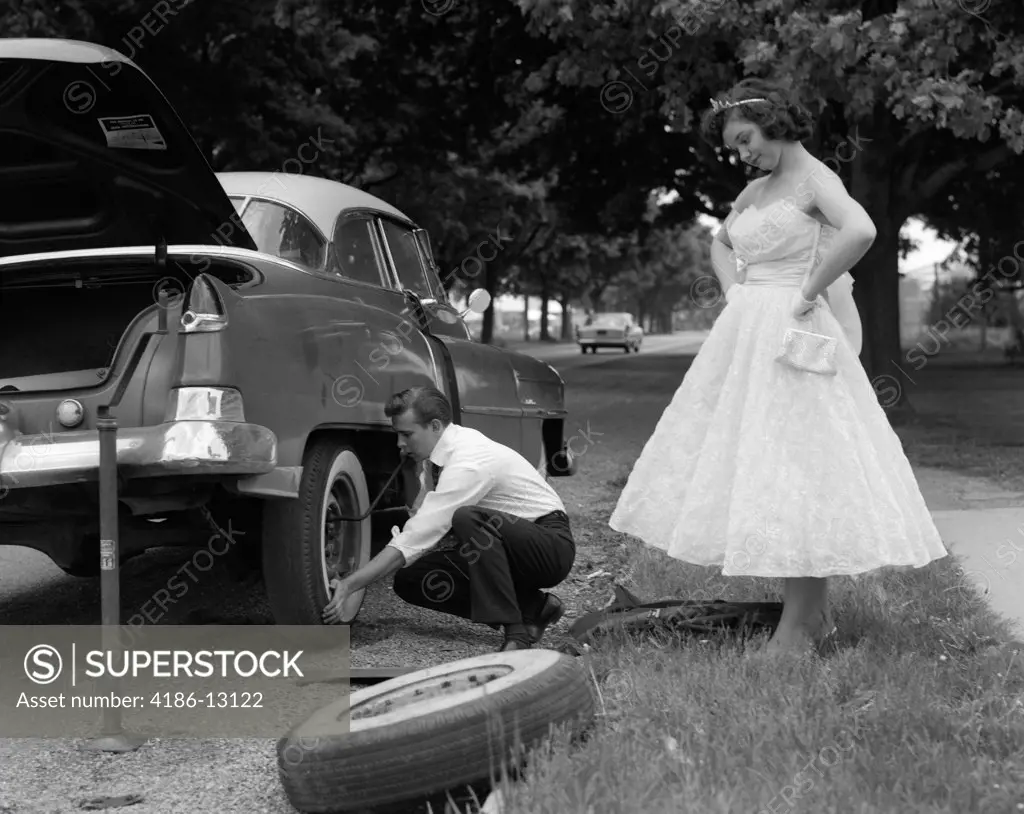 1950S 1960S Teenage Boy Crouching Down Changing Flat Tire Prom Date Watching With Hands On Hips