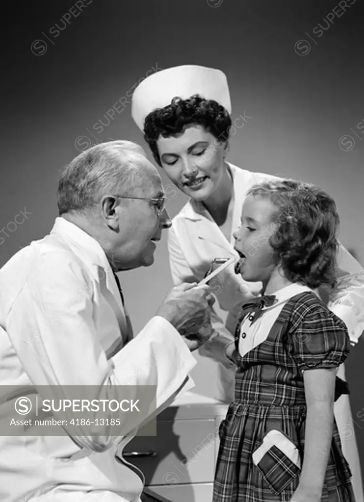 1950S Doctor Examining Mouth Of Little Girl With Tongue Depressor While Nurse Is Looking On