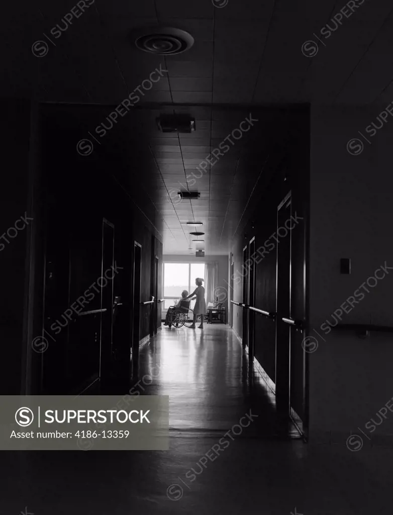 1960S 1970S Nurse Pushing Elderly Female Patient Down Dark Hospital Hallway In Wheelchair