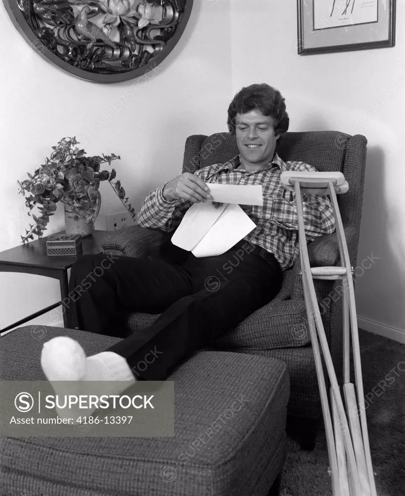1970S Young Man In Arm Chair With One Leg Propped Up Crutches Nearby Reading A Letter Indoor