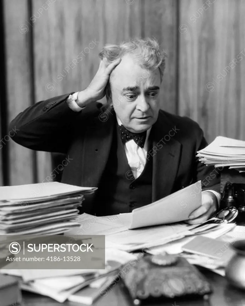 1930S Elderly Businessman Sitting At Desk Among Stacks Of Papers With Hand In Hair & Flustered Expression