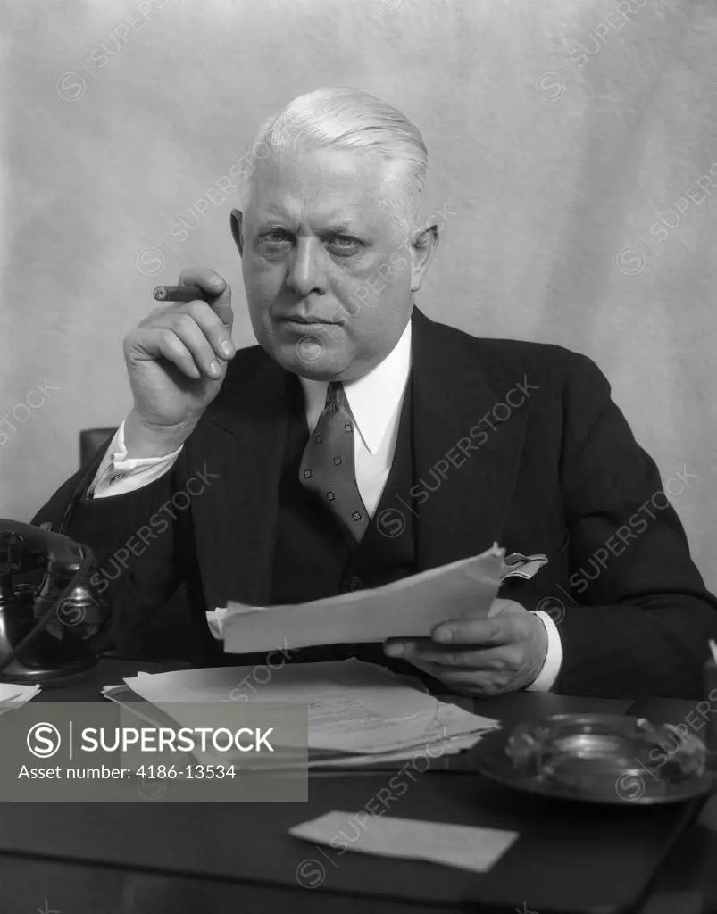 1930S Man In Office Sitting At Desk Holding Papers Smoking A Cigar