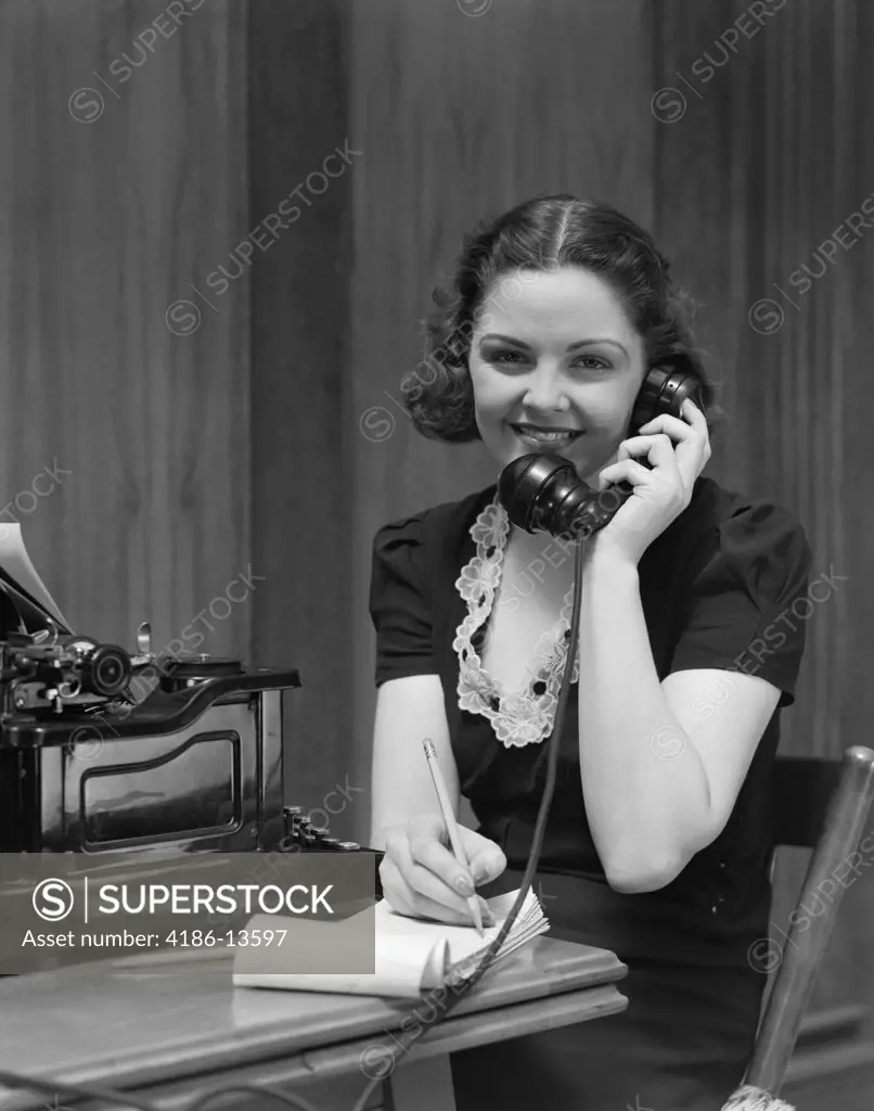 1930S Smiling Young Woman Receptionist Secretary Sitting At Desk In Office Talking On Telephone