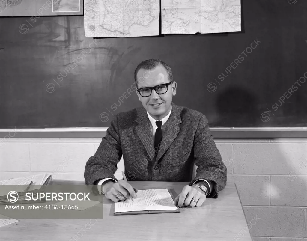 1960S Man Teacher At Desk In Classroom Pencil Paper In Front Blackboard Background Smiling At Camera