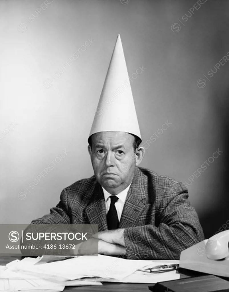 1960S Unhappy Businessman Behind Desk Wearing Dunce Cap Looking At Camera