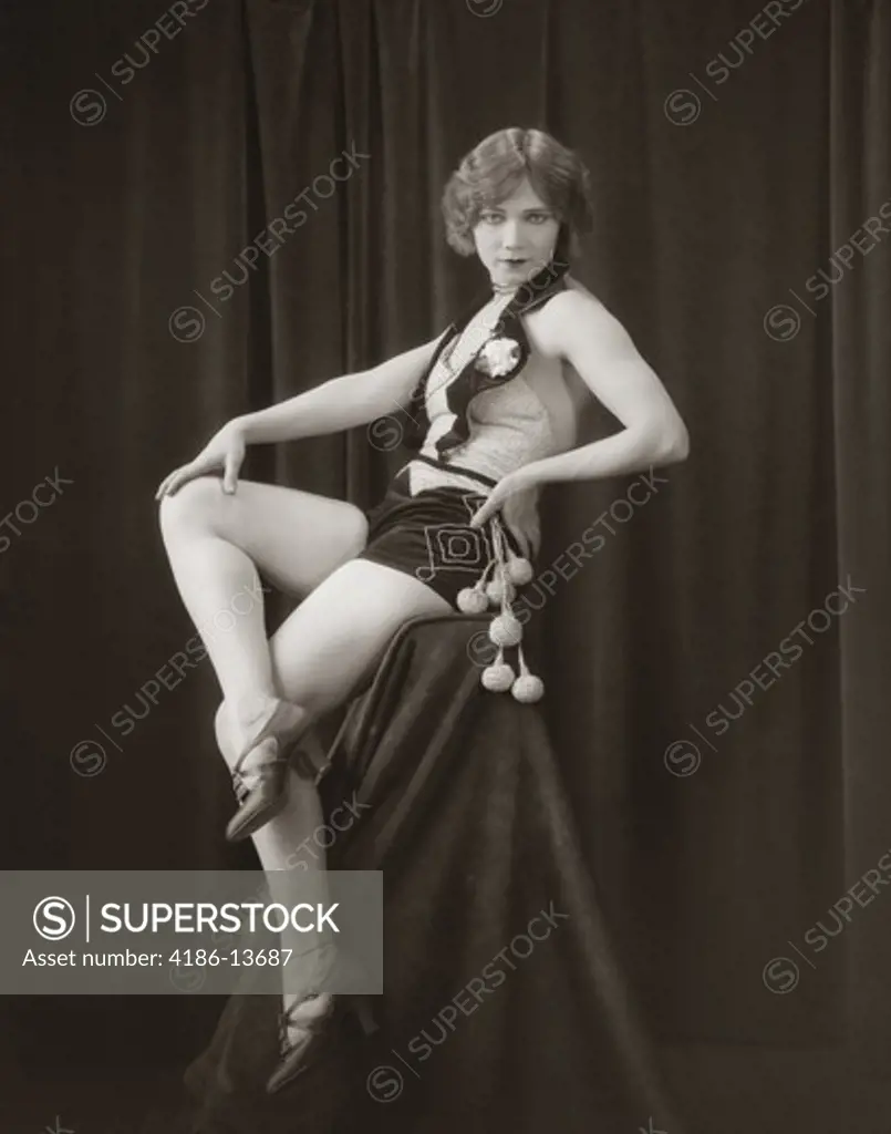 1920S Side View Of Flapper Woman In Velvet Hot Pants & Sequined Vest Sitting On Pedestal With Legs Crossed Looking Toward Camera