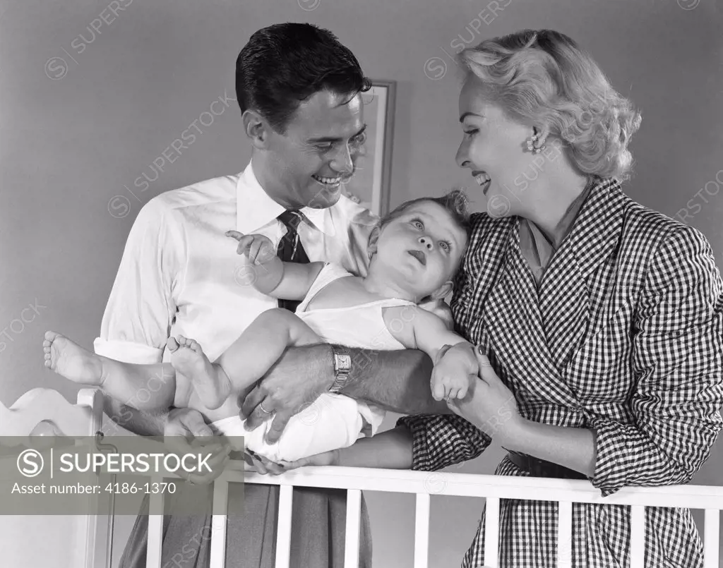 1950S Smiling Parents Picking Baby Up Out Of Crib