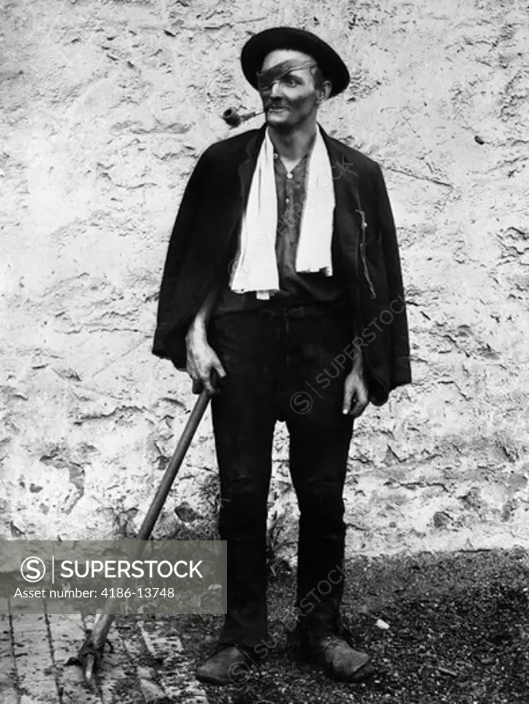 1890S 1900S Turn Of The Century Man With Eye Patch & Pipe In Mouth Wearing Hat & Scarf Around Neck Holding Pick At Side