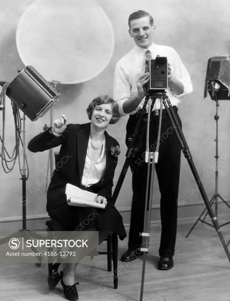1920S Photographer Standing Behind Camera In Studio With Assistant Sitting Next To Him Holding Paperwork