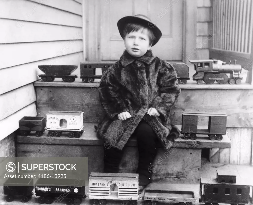 1880S 1890S 1900S Boy Wearing Fur Coat Sitting On Steps Surrounded By Toy Railroad Trains Looking At Camera