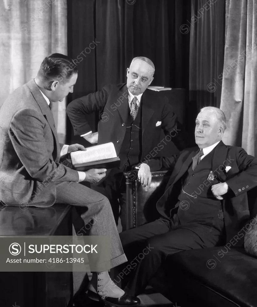 1940S Group Of Three Businessmen Gathered Around Having Discussion One Referring To Book