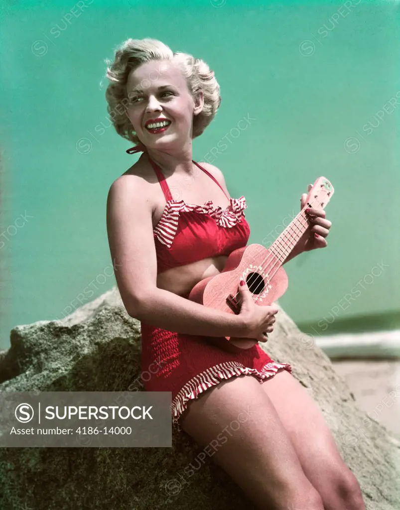 1940S 1950S Smiling Blond Woman Wearing Red Bikini Bathing Suit Leaning On Rock At Beach Playing Ukulele