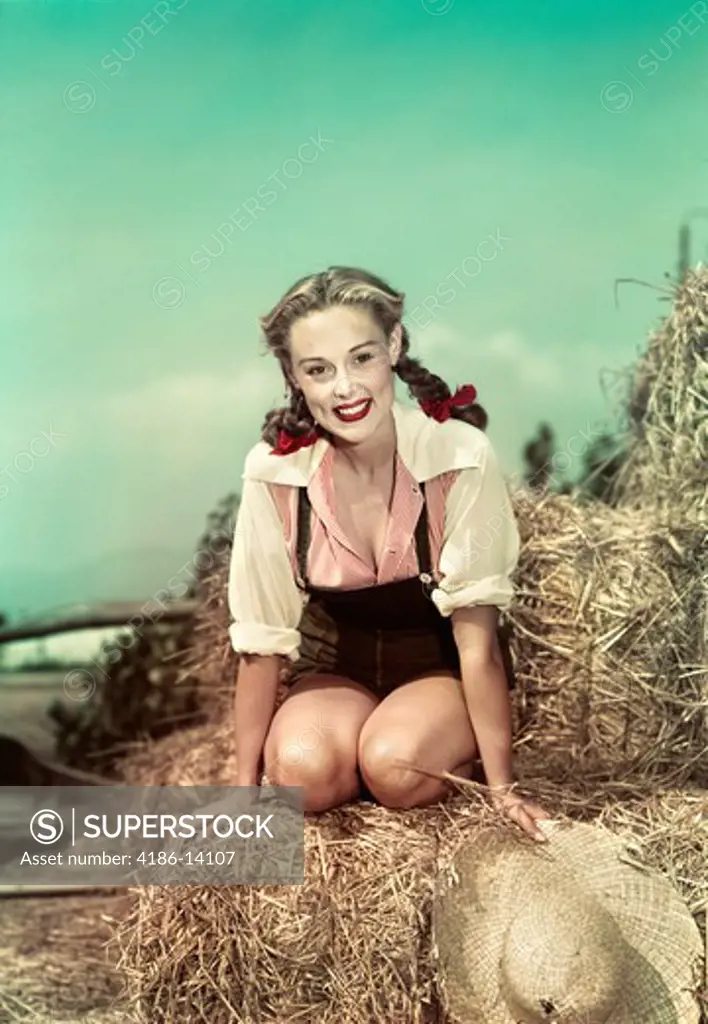 1940S 1950S Smiling Woman Hair In Braids Wearing Overalls Sitting On Hay Stack Holding Straw Hat