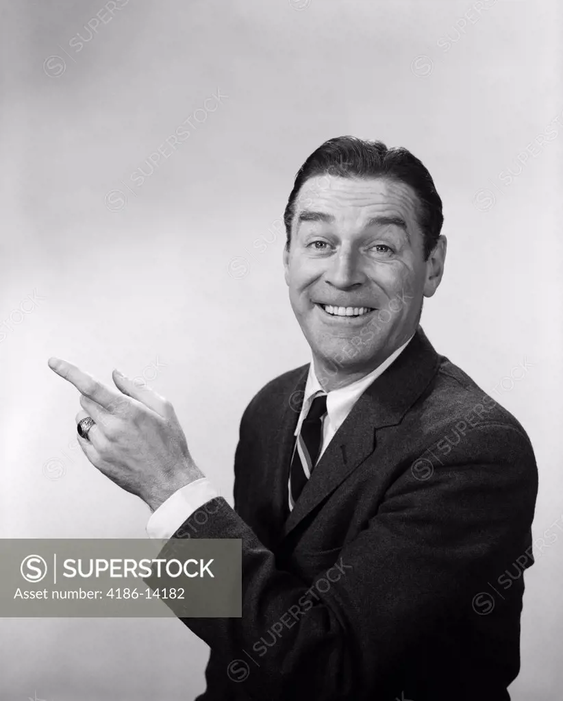 1960S Smiling Happy Man Business Spokesperson In Suit And Tie Looking At Camera Pointing Hand And Index Finger