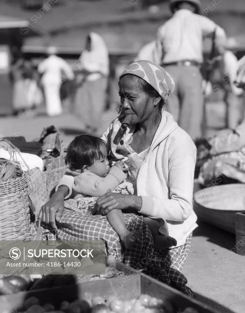 1920S 1930S Mother Sitting Holding Baby Child In Market Place Woman Smoking Cigar Cheroot Baguio Philippines