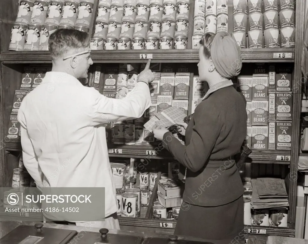 1940S Back View Of Grocer Pointing Out Prices Of Canned Goods To Woman Holding Ration Book