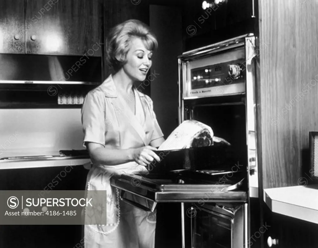 1960S Woman Wearing Apron In Kitchen Putting Roast Into The Oven