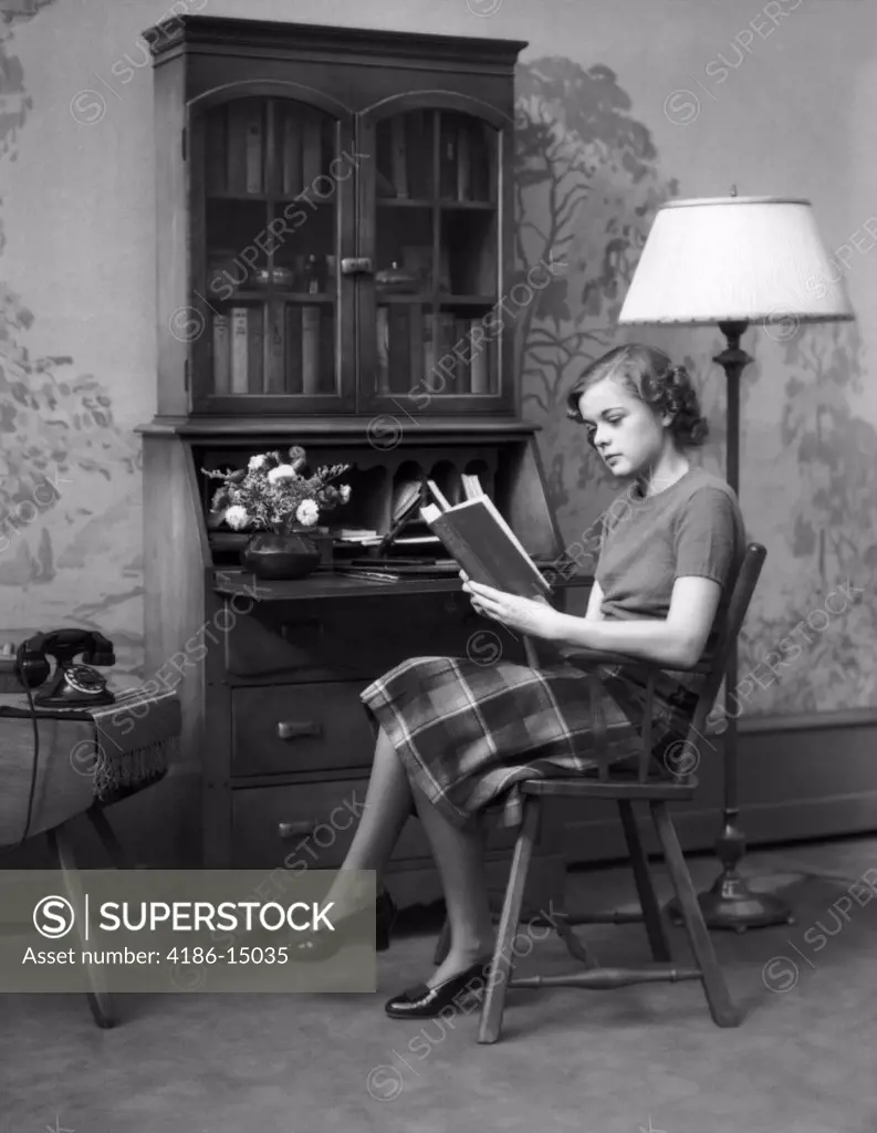 1930S 1940S Young Woman Teenage Sitting In A Chair Reading A Book By A Maple Wood Secretary Desk In Peaceful Home Living Room