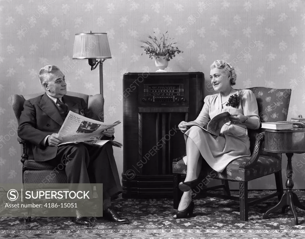 1940S Senior Couple Sitting In Living Room Reading Newspaper And Magazine Listening To Radio