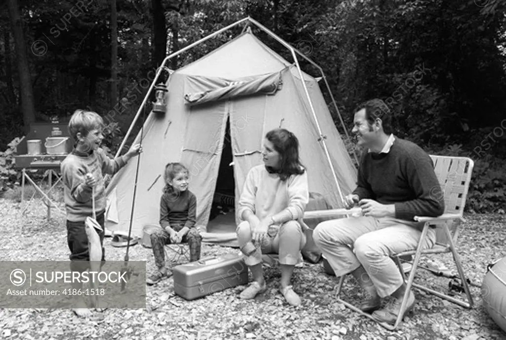 1970S Family Of Four Sitting In Front Of Tent In Woods Son Holding Fishing Pole In One Hand & Big Fish In Other
