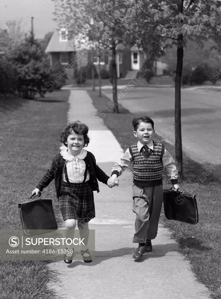 Boy And Girl Carrying Book Bags Walking Together Hand In Hand On Sidewalk To Or From School Outdoor