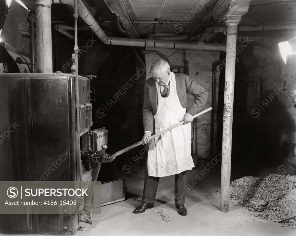 1940S Elderly Man In Basement Wearing Apron Shoveling Coal Into Furnace