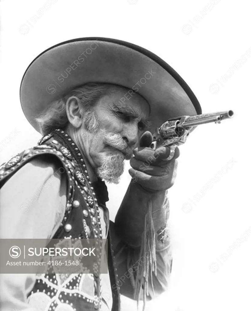 Senior Bearded Cowboy Pointing A Revolver At The Camera Dressed In Fringed Glove Beaded Vest Wide Brimmed Hat