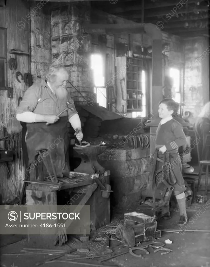 1930S Elderly Blacksmith With Hammer At Anvil As Young Boy Holding Harness Looks On Smiling