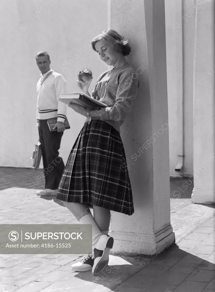 1950S 1960S Teen School Girl Plaid Skirt Leaning Against Column Eating Apple Reading Book As Boy Walks By In Background