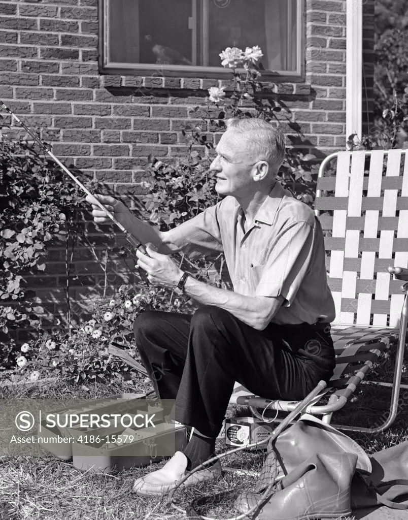 1960S Elderly Man Seated On Lawn Chair Fiddling With Fishing Rod With Tackle Box Net & Rubber Boots At Feet