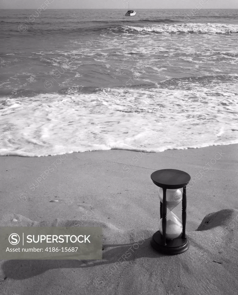 1960S Set-Up Of Hourglass On Ledge Of Sand With Waves Washing Up On Shore & Boat Coming In Background