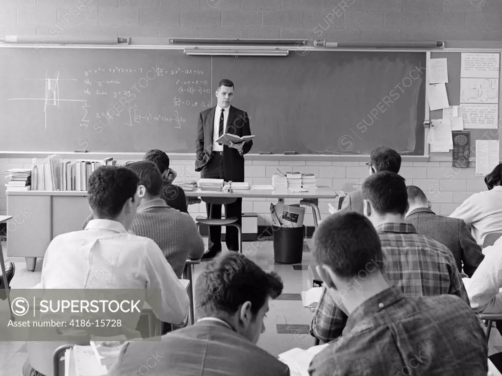 1960S Back View Of Classroom With Teacher Standing Behind Desk With Book In Hand Explaining Graphed Equation On Blackboard