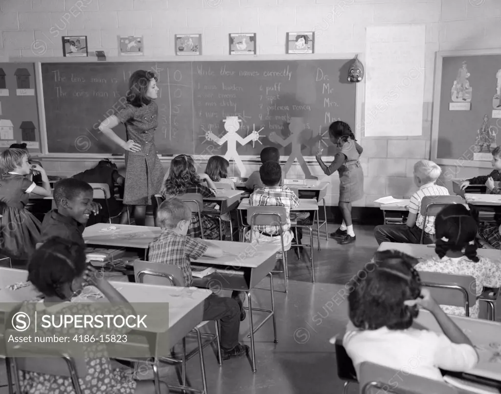 1960S Back View Of Grade School Classroom With Teacher At Front Watching Student Working On Blackboard