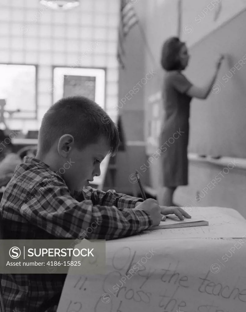 1960S Side View Of Grade School Boy At Desk Writing With Teacher Writing On Blackboard In Soft Focus In Background