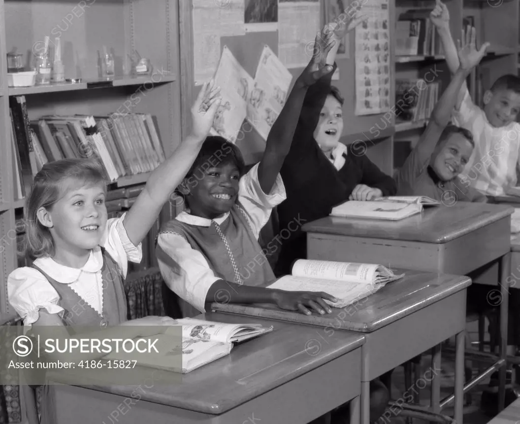1960S Group Of Grade School Children At Row Of Desks Eagerly Raising Hands