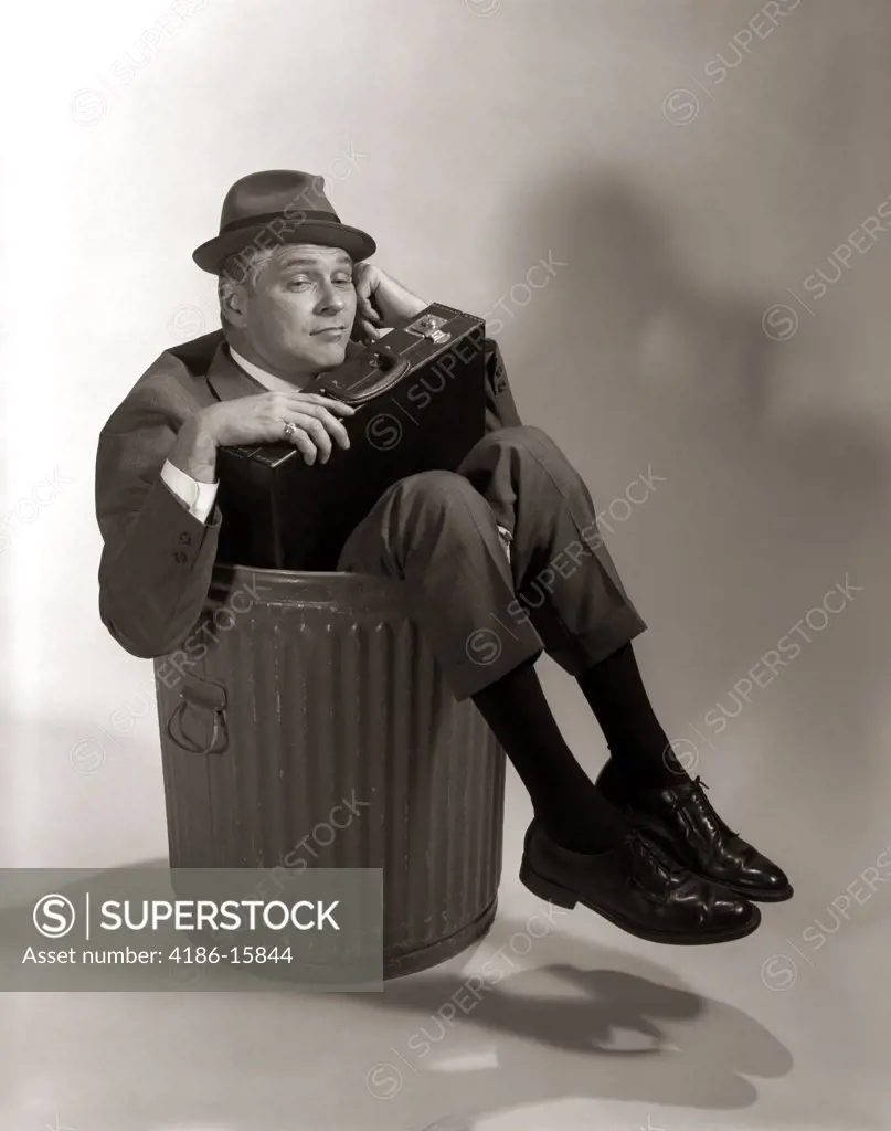 1960S Symbolic Businessman In Suit And Hat Holding Briefcase Dumped Fired Made Redundant Laid Off Unemployed Sitting In Trash Can