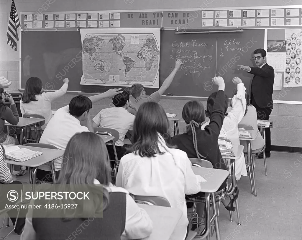 1970S Man Teacher Pointing To Raised Hand Teen Classroom Students Seen From Behind Look To Chalkboard World Map History Class