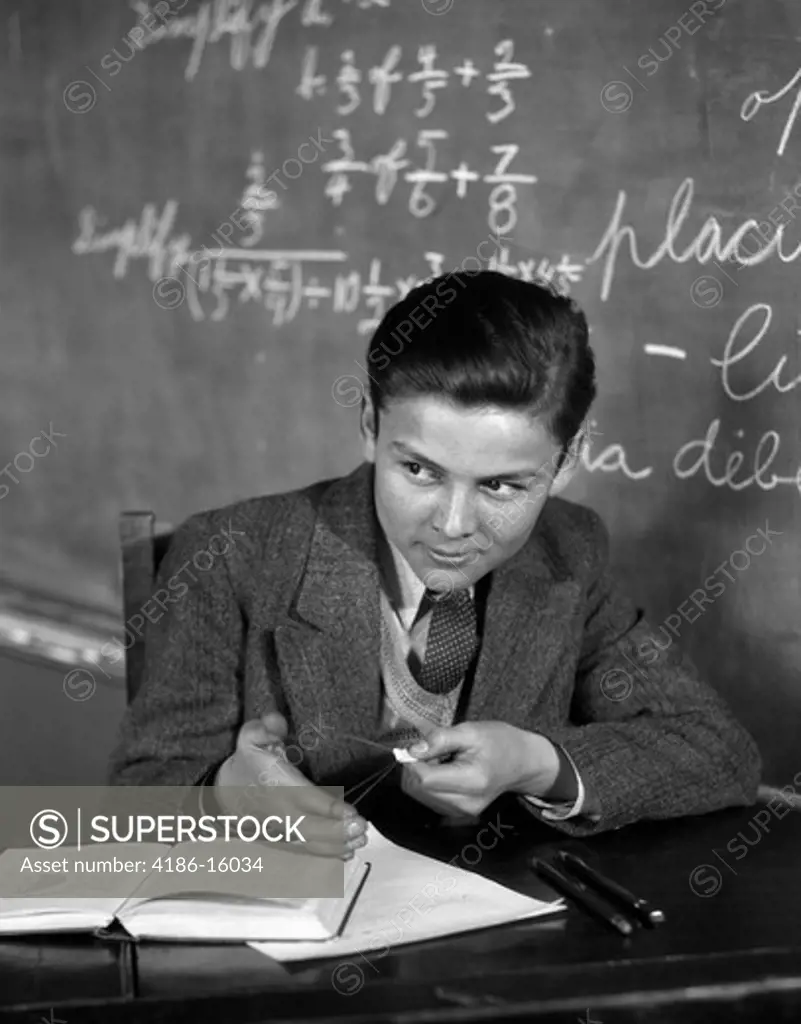 1920S 1930S Boy At Desk In Classroom In Front Of Blackboard Shooting Paper Wad With Rubber Band