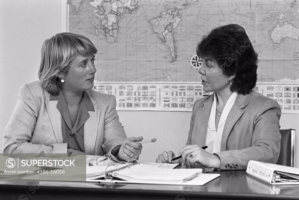 1980S Pair Of Female Teachers Having Discussion Notebooks Spread Out On Desk In Front Of Them World Map On Wall In Background