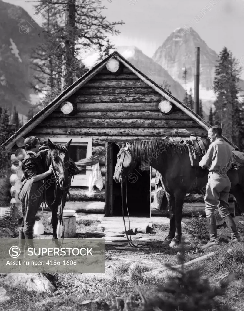 1920S 1930S Couple In Front Of Log Cabin Standing By Horses With Saddles Both Wearing Jodhpurs Assiniboine Canada