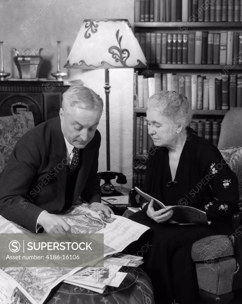 1930S Elderly Couple Man Woman Read Maps Books Travel Brochures Sit In Living Room Book Shelves
