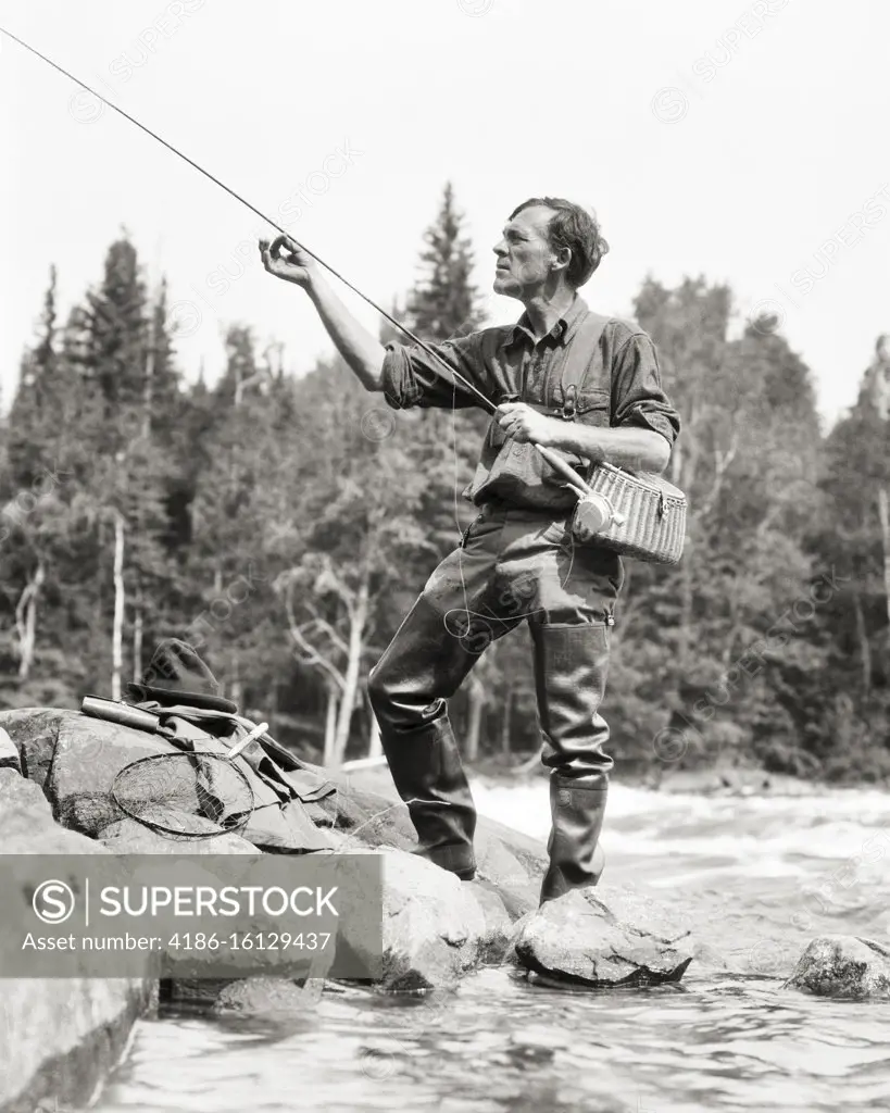 1920s FISHERMAN ADJUSTING HIS FISHING ROD BEFORE CASTING IN STREAM IN CANADA  - SuperStock