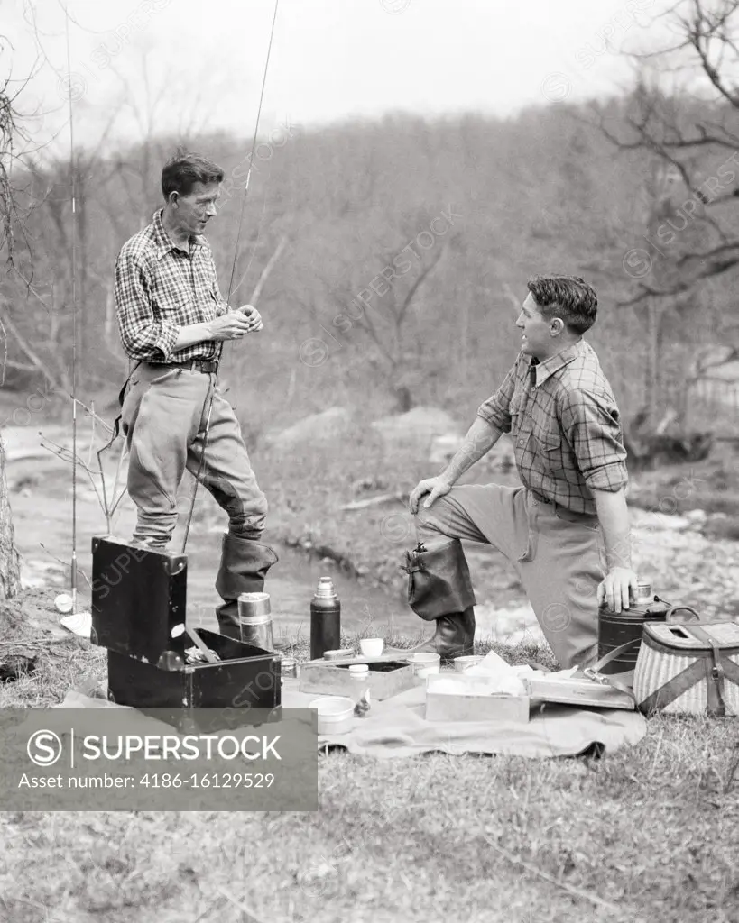 1930s TWO MEN WITH FISHING GEAR RODS CREEL WADERS TACKLE BOX HAVING PICNIC  LUNCH COFFEE FROM THERMOS - SuperStock