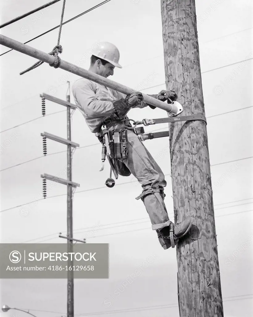 1960s ELECTRIC LINEMAN ON UTILITY POLE SECURED WITH SAFETY BELT AND GAFFS  OR SPURS ON HIS FEET WEARING A HARD HAT AND TOOL BELT - SuperStock