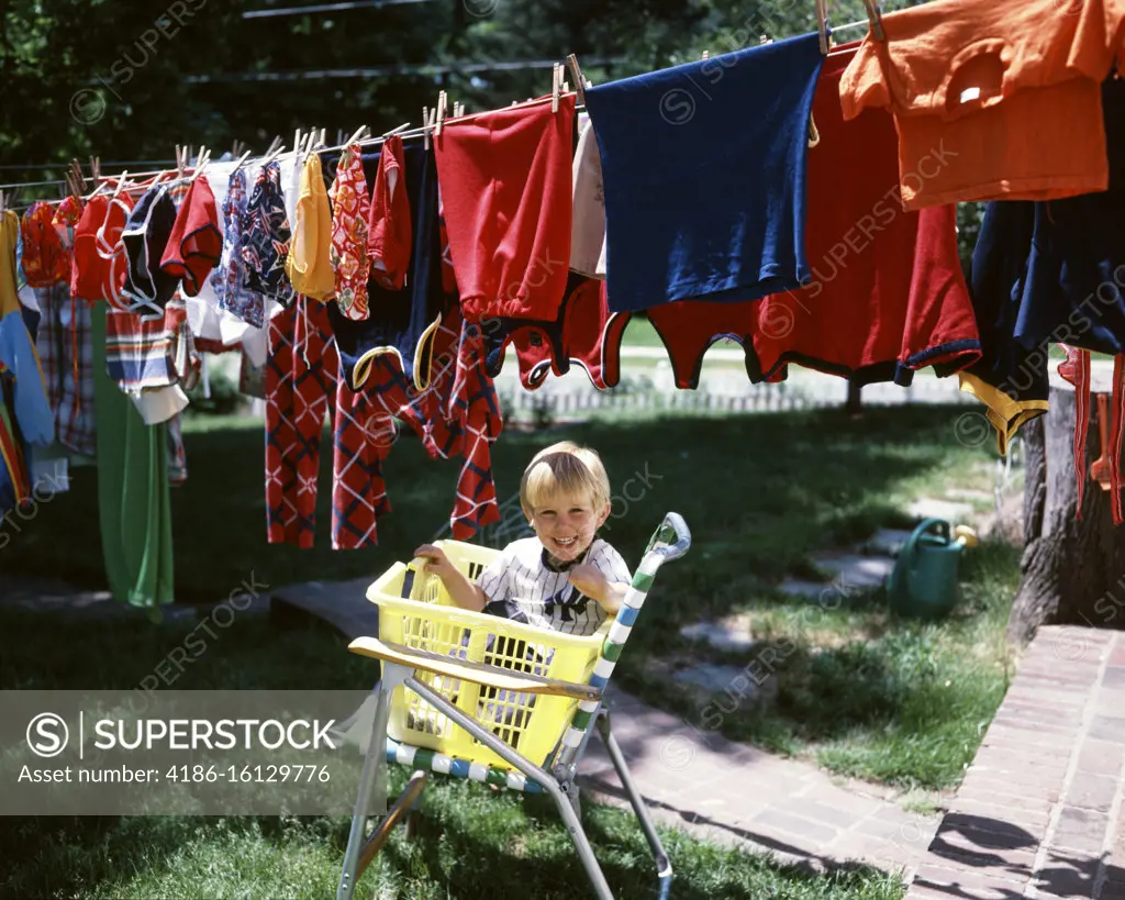 1970s LAUGHING BLOND BOY LOOKING AT CAMERA SITTING IN YELLOW LAUNDRY BASKET UNDER CLOTHESLINE FULL OF COLORFUL WASHED CLOTHES
