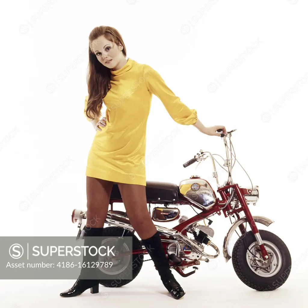 1960s SEXY BRUNETTE WOMAN WEARING A YELLOW MINIDRESS HIGH BLACK BOOTS LOOKING AT CAMERA STANDING BESIDE A SMALL MOTOR BIKE