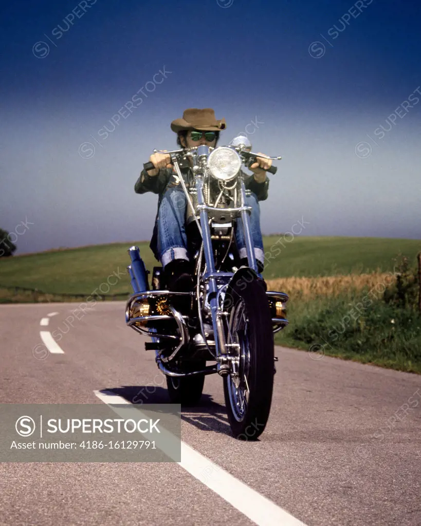 1970s MAN WEARING HAT SUNGLASSES ROLLED CUFF BLUE JEANS LOOKING AT CAMERA RIDING CUSTOM BUILT CHOPPER MOTORCYCLE ON HIGHWAY