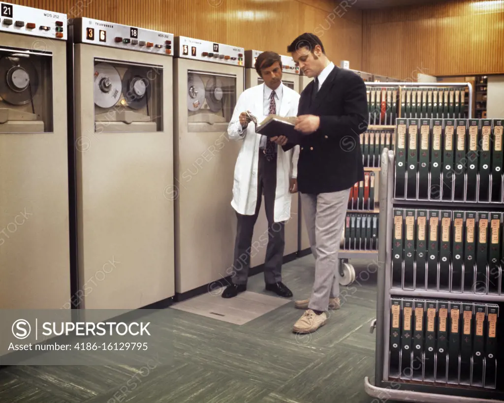 1980s TWO ANONYMOUS MEN MANAGER AND TECHNICIAN STANDING TOGETHER CHECKING A MANUAL BESIDE COMPUTER MAINFRAME TAPE STORAGE SILOS
