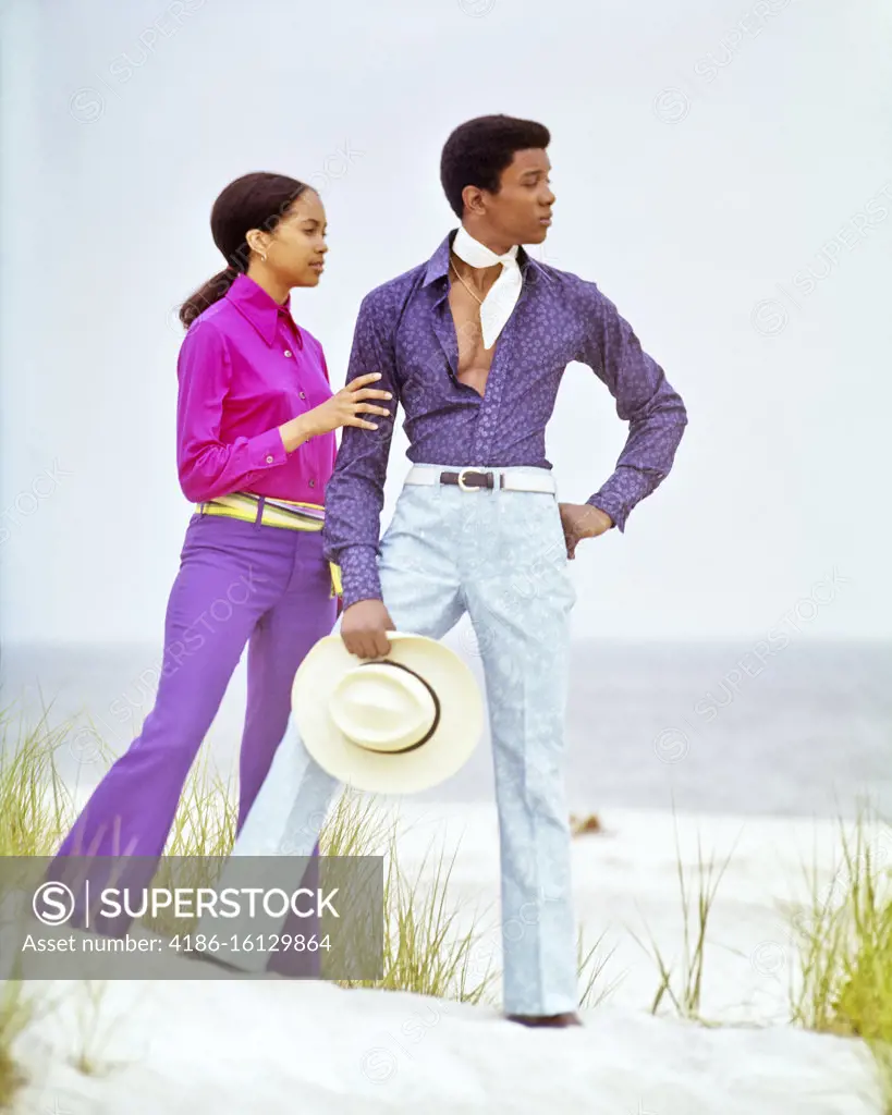 1970s YOUNG ADULT AFRICAN AMERICAN COUPLE STANDING ON BEACH DUNE WEARING COLORFUL SHIRTS BELL BOTTOM PANTS LONG ISLAND NYC USA